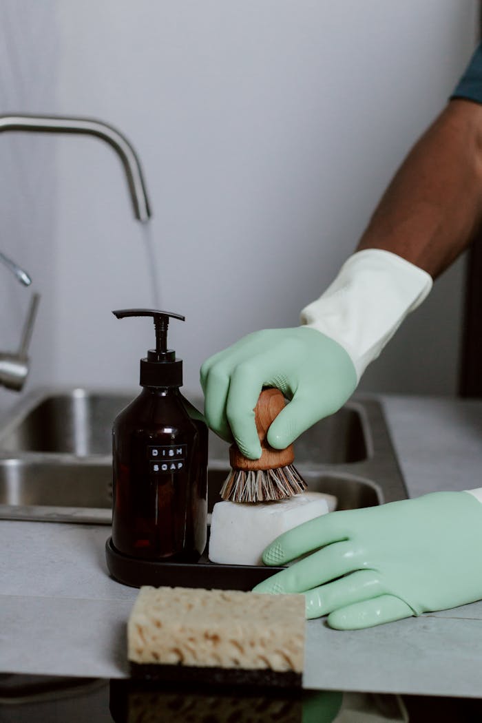 Close-up of hands in green gloves washing dishes with dish soap and brush in a kitchen sink.